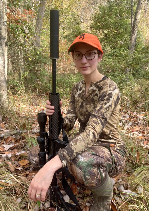 Girl wearing First Lite Women's Wick Quarter Zip kneeling in the woods with a rifle.