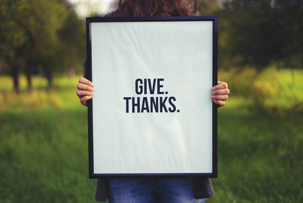 Person holding a white sign outside that says give thanks in black letters. 