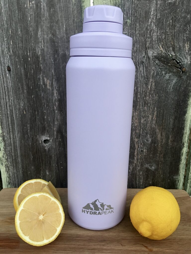 A light purple water bottle sitting on a wooden board with a fence in the background and lemons on either side. Hydrapeak water bottle review. 
