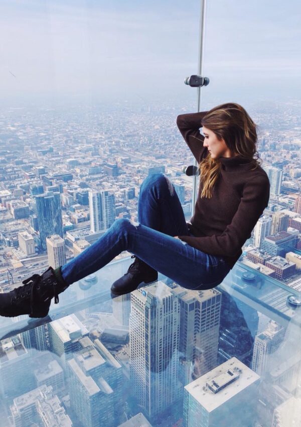 Girl wearing a brown turtle neck and ripped jeans and boots sitting on the edge of the Ledge at the Chicago Skydeck