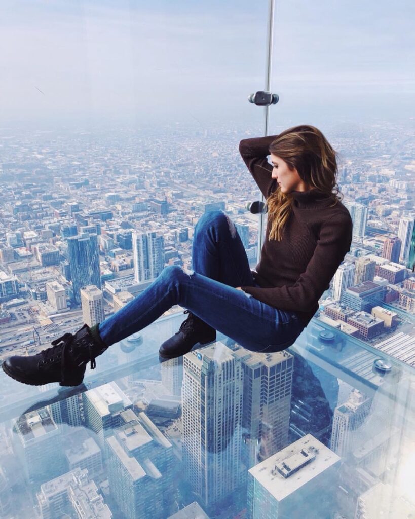 Girl wearing a brown turtle neck and ripped jeans and boots sitting on the edge of the Ledge at the Chicago Skydeck