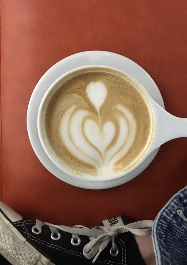 A latte sitting on a reddish orange seat with a girls converse shoes in the shot. A picture from Coat Check Coffee during a weekend in Indianapolis.