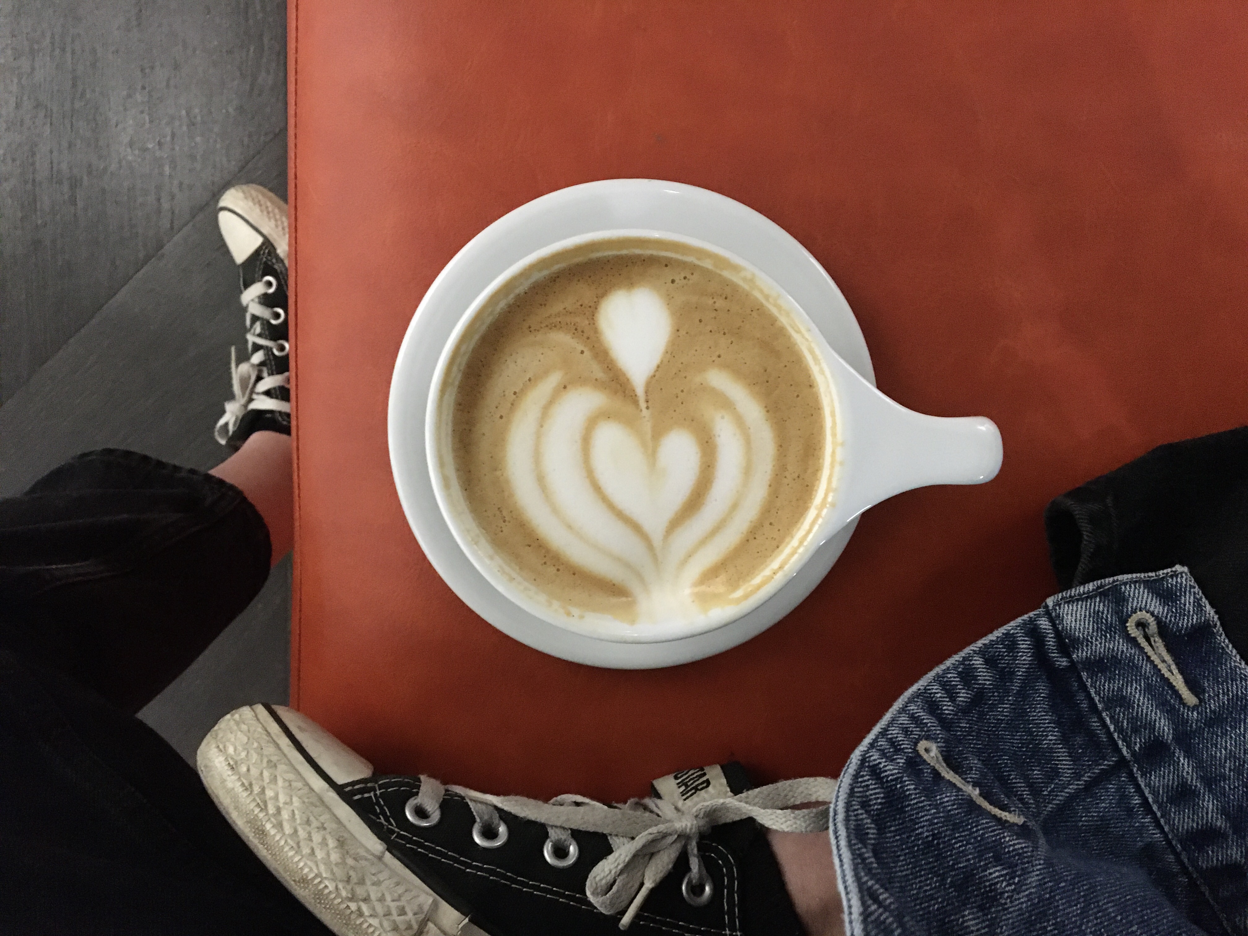 A latte sitting on a reddish orange seat with a girls converse shoes in the shot. A picture from Coat Check Coffee during a weekend in Indianapolis. 