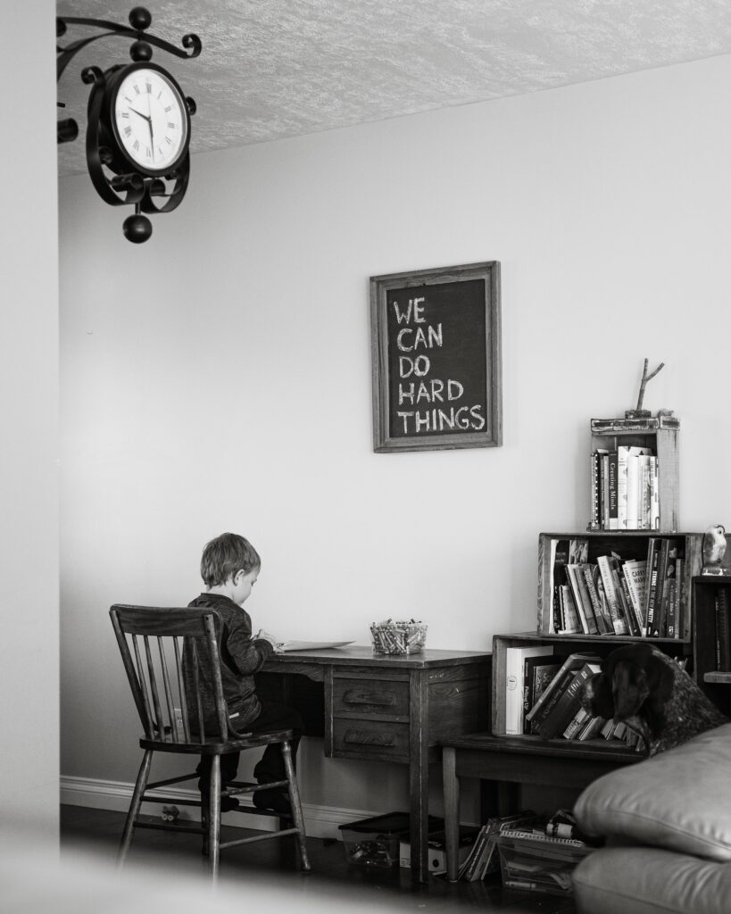 A little boy sitting at a desk doing school work with a sign that says we can do hard things. 